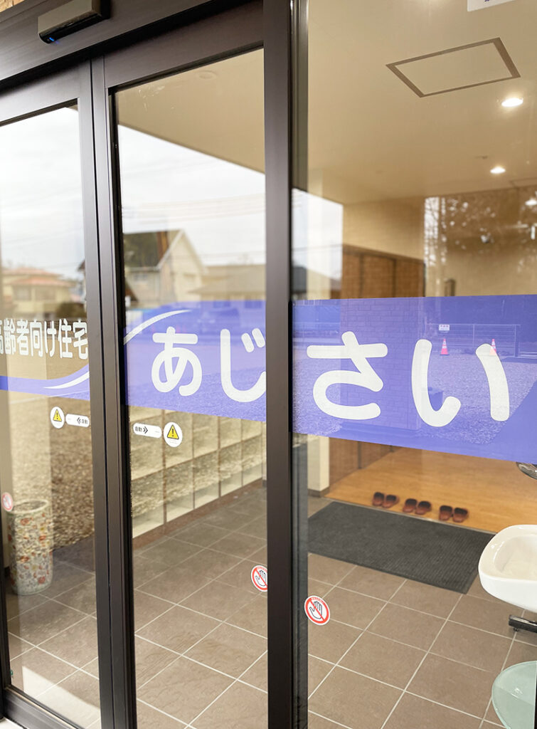 介護施設の玄関。ガラス戸に施設名が表示され、エントランスマット・靴箱が見える。